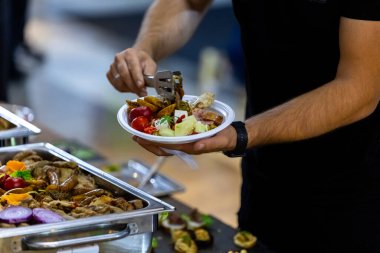 Bir insan açık büfede ızgara sebze ve et servis etmek için maşa kullanır. Atmosfer canlı, çeşitli yiyecek seçenekleri ve güneşli havayla dolu..