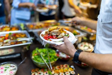 Canlı bir ortamda, bir insan et ve salata da dahil olmak üzere farklı yiyeceklerle dolu bir tabağı tutarken, bir dizi renkli açık büfe yemekleri ve yemeklerin tadını çıkaran insanlarla çevrelenir..