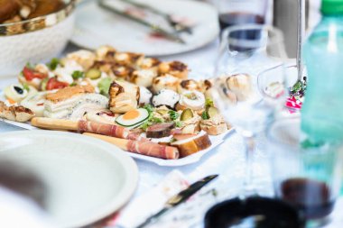 Çeşitli malzemelerin yer aldığı kanepeler beyaz bir tabakta zarif bir şekilde sunuluyor. Bardaklar ve tabaklarla çevrili, samimi bir yemek düzeninde..