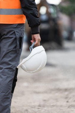 Turuncu güvenlik yeleği giymiş bir inşaat işçisi, bir elinde beyaz bir şapka olan, engebeli bir inşaat sahasında yürür ve ilerideki görevlere odaklanır..