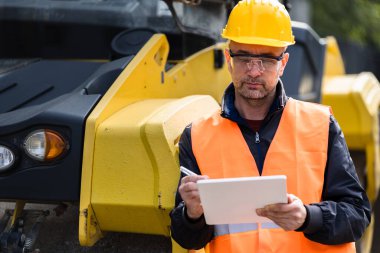 Parlak sarı şapka ve güvenlik yeleği giyen bir inşaat işçisi, şehir merkezinde ağır makinelerin yanında dururken not alıyor..