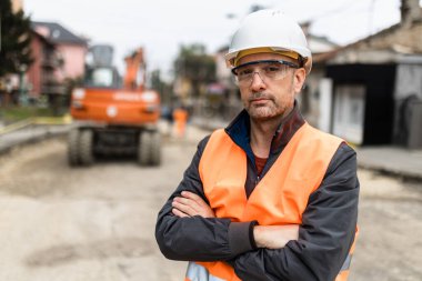 Bir inşaat yöneticisi emniyet yeleği ve sert bir şapka takarak güvenle ayakta duruyor, yakınlarda makineler bulunan kentsel bir çevrede yol çalışmalarını denetlerken,.
