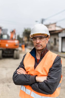 Turuncu yelek giyen ve gözlüklü bir şapka takan bir inşaat işçisi, kentsel bir iş yerinde kendine güvenen bir tavır sergiliyor ve kameraya doğrudan bakıyor..
