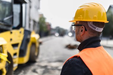 Güvenlik teçhizatı kullanan bir inşaat işçisi, şehir caddesinde çalışan ve gün ışığında projenin güvenliğini ve verimliliğini sağlayan büyük makineleri gözlemler..