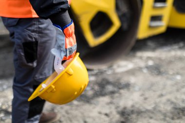 Koruyucu giysili bir inşaat işçisi açık havada elinde sarı bir şapkayla beklerken açık mavi gökyüzünün altında büyük sarı bir yol silindiri çalışıyor..