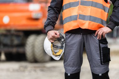 İnşaat işçisi, inşaat sahasında, emniyet teçhizatı takmış ve sıkı bir şapka takmış bir şekilde, inşaat işindeki güvenliğin önemini vurguluyor..