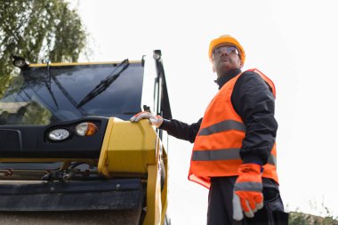 Turuncu güvenlik yeleği giyen ve kasketli bir inşaat işçisi, bir şantiyedeki operasyonları denetlerken ağır bir mekanizmayı inceliyor..