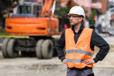 Kask ve yansıtıcı yelek giyen bir inşaat işçisi, elleri kalçasında, açık gökyüzünün altındaki işlek bir inşaat alanında çalışan makineleri gözlemler..