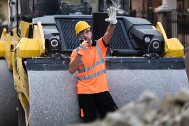 Parlak turuncu yelek ve kasklı bir inşaat işçisi, geçen araçlara yol silindirinin yanında dururken işaret veriyor. Kentsel zemin etkinliği artırır.