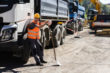 Sert şapka ve yansıtıcı yelek giyen bir inşaat işçisi, gün ışığında yoğun bir şantiyede kamyonlar ve ağır makinelerle çevrili bir küreğin üzerinde dinleniyor..