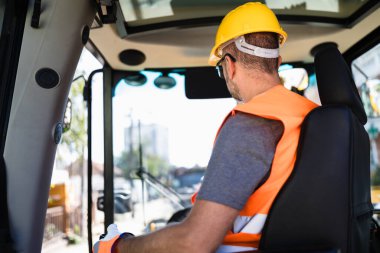 Bir inşaat işçisi gündüz vakti ağır makineleri sürmeye, şehir ortamında güvenlik ekipmanları giymeye odaklanır. Bu da yoğun bir iş günü olduğunu gösterir..