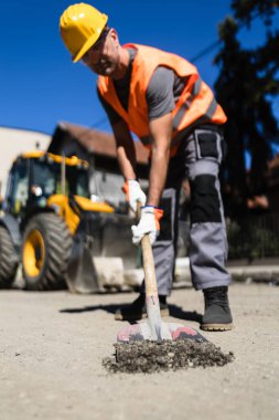 Turuncu yelek ve sert şapkalı bir inşaat işçisi kaldırımı tamir etmek için kürek kullanıyor. Arka planda, etkin bir çalışma alanını gösteren ağır makineler görülüyor.