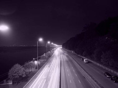 A stunning long-exposure nighttime photograph of a highway illuminated by streetlights, capturing light trails of passing vehicles. The city skyline glows in the distance, reflecting off the river.  clipart