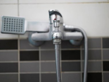 Eski bir krom banyo musluğunun yakın çekimi. Duş başlığı takılı, fayanslı bir banyo duvarına monte edilmiş. Yıllanmış metal yüzeyi kırsal ve endüstriyel bir estetik verir.