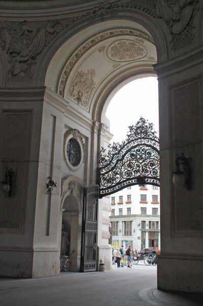 Элегантная историческая арка со сложными деталями в стиле барокко и украшенными коваными железными воротами, ведущими на оживленную европейскую улицу. 