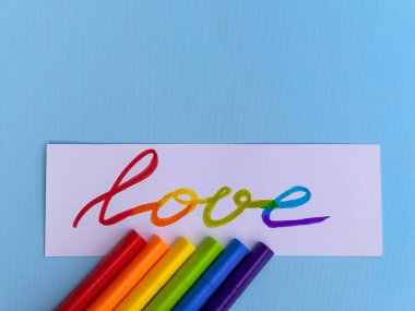 Aşk ve mavi arkaplan yazılı beyaz bir kartın başvurusu. Gökkuşağı lgbt bayrağının renklerindeki renkli keçeli kalemlerin yanında