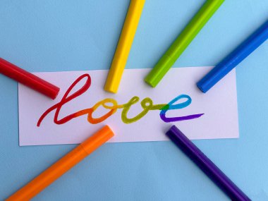 Aşk ve mavi arkaplan yazılı beyaz bir kartın başvurusu. Gökkuşağı lgbt bayrağının renklerindeki renkli keçeli kalemlerin yanında