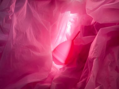 Yönlendirilmiş bir ışık demeti altında pembe kumaş kıvrımlı adet bardağı. İlahi ışıltı, ana nesneye odaklan, renk tonlarını kapat. Bloglar, makaleler, adet görselleri için