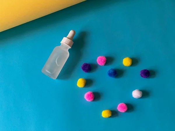 stock image Serum in a glass dropper bottle on a blue and yellow background with a multicoloured pom-poms folded in the shape of a heart. Skincare routine. 