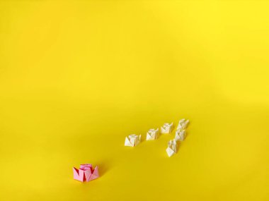 Origami tekneleri. Bir grup beyaz kâğıt gemi ve sarı arka planda bir pembe kağıt tekne. Takım çalışması ve liderlik kavramı.