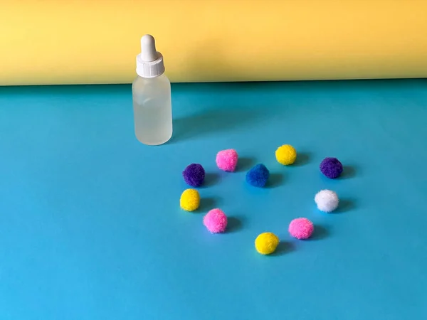 Serum in a glass dropper bottle on a blue and yellow background with a multicoloured pom-poms folded in the shape of a heart. Skincare routine.