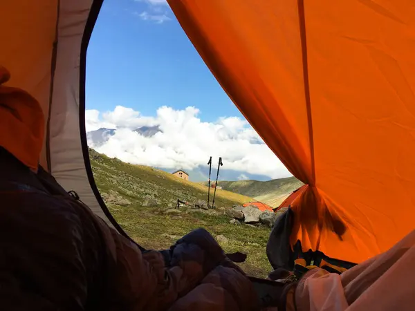 Panoramic view from the window of the tourist tent in orange color. Trekking sticks stand against the background of mountains, fluffy clouds and blue sky