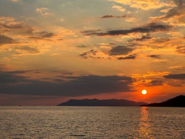 Gün batımı dağların arkasında, denizin önünde batar. Gökkuşağının tüm renkleriyle aydınlatılan bulutlar