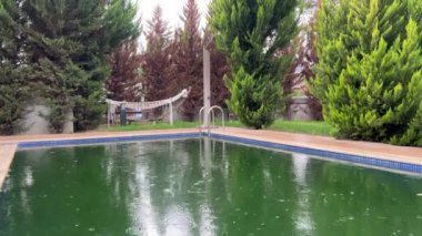 Büyük yağmur damlaları havuza düşer ve suda daireler çizer. Turizm sezonunun sonu, terk edilmiş villa, yeşil havuzda su.