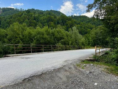 Arka planda yemyeşil tepeler olan sessiz bir köprüyü geçen ıssız bir yol..