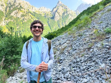 Arkasında dağlar olan kayalık bir yamaçta duran gülümseyen bir yürüyüşçü..