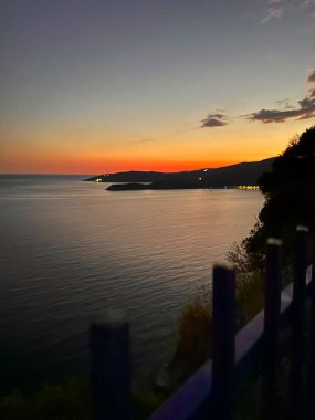 Sakin deniz üzerinde gün batımı siluetli kıyı şeridi ve renkli gökyüzü.