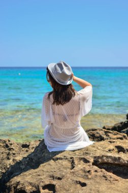 Deniz kıyısında şapkalı genç bir kadın. Beyaz elbiseli ve şapkalı bir kadın okyanusta oturuyor. Kız deniz kıyısındaki bir kayanın üzerinde oturuyor. Bir kadın deniz kıyısına bakıyor..