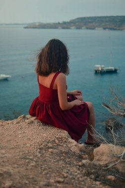 Deniz kıyısındaki bir kadın kayanın üzerinde oturur. Deniz kenarında elbise giymiş bir kadın. Güzel pozlar verir. Rüzgâr saçlarını savurur. Fotoğraf için güzel bir yer..