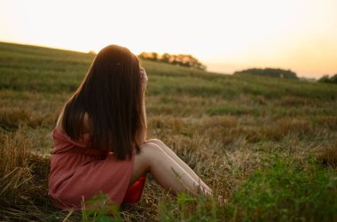 Elbiseli bir kadın gün batımında bir tarlada oturur ve uzaklara bakar. Tarlada uzun saçlı bir kız. Güzel bir yaz elbisesi. Gün batımında manzara. Güzel saçlı, kendine güvenen bir kadın.