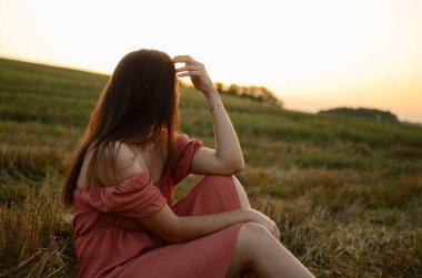 Elbiseli bir kadın gün batımında bir tarlada oturur ve uzaklara bakar. Tarlada uzun saçlı bir kız. Güzel bir yaz elbisesi. Gün batımında manzara. Güzel saçlı, kendine güvenen bir kadın.