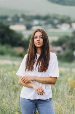Bir kadın portresi. Güzel bir kız tarlada yürüyor. Beyaz tişörtlü bir kadın. Güzel doğa. Makyajsız doğal güzellik. Güzel saçlı esmer.