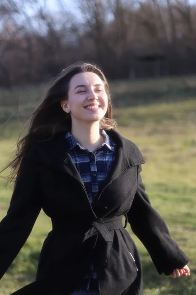 a young brunette girl in a black sweater and black jacket stands in the park in spring.Portrait of a young girl in nature.Vintage retro photo.Photo in motion.Long hair.Wind blowing on her hair.Extraordinary interesting photo.Lady in black.Sunlight.