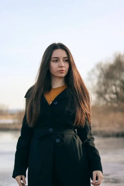 beautiful girl in a long coat on the background of the river.Portrait of a woman.Portrait of a brunette girl.Woman in black clothes.A woman with long hair in a black coat.Beautiful face.The brunette stands near the water.The girl looks to the side.
