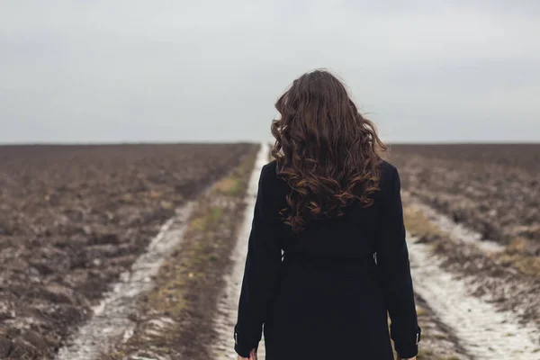 a girl in a field with a black coat.A girl in a black coat with long curly hair walks along a dirt road.A lonely woman walks in the field.A girl walks along the road.Retro vintage photo.Gloomy atmosphere.Loneliness,thoughts and dreams.