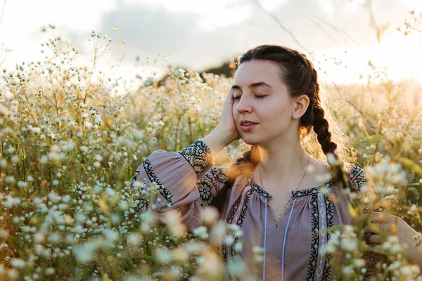 Ukrainian woman in an embroidered shirt sits in a field of daisies at sunset.dreaming woman.pensive happy woman.self-confident girl.patriotism.woman on a flowering meadow.a tender girl.Loneliness.girl with pigtails in nature.portrait in the meadow