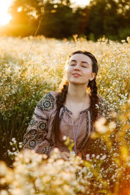 Nakışlı gömlekli Ukraynalı kadın gün batımında papatya tarlasında oturur. Dalgın mutlu kadın. Kendine güvenen kız. Vatansever kadın..