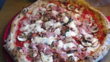 Kırmızı bir tabakta, ahşap bir masanın üstünde lezzetli dokuları ve taze malzemelerin altını çizerek servis edilen jambon ve mantarlı pizzanın aşırı yakın çekim ve hareketli görüntüsü.. 