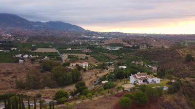 İnsansız hava aracı, Mijas, Mlaga, İspanya 'da yeşil bir arazide sağa sola doğru hareket ediyor. Dağlar, havuzlu büyük beyaz bir villa, ana yol ve bulutlu bir gökyüzünün altında uzak bir tepe..