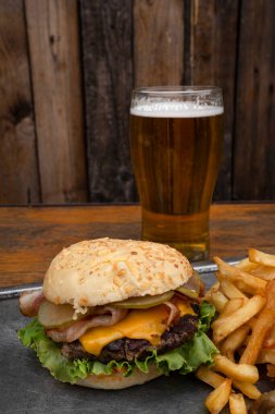 Lezzetli hamburger. Çedar peyniri, salatalık ve kızarmış domuz pastırmalı hamburger, patates kızartması ve ahşap bir arka plan.. 