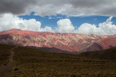 Jeoloji. Renkli Hornocal dağına uzanan sarı çayır boyunca uzanan kirli bir yol dramatik bulutlu bir gökyüzünün altında.
