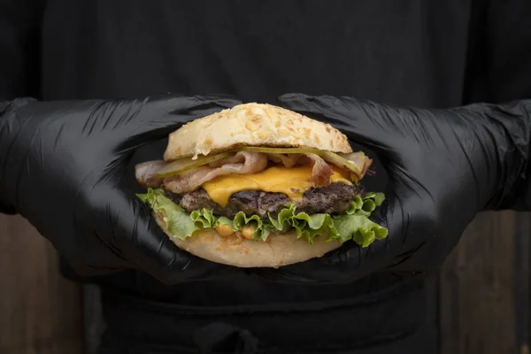 Chef wearing gloves holding a burger made with bread, sliced cucumber, lettuce, meat, bacon and cheddar cheese.