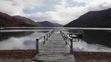 Dağlar ve ormanla çevrili göldeki ahşap rıhtım manzarası. Gökyüzü ve bulutlar su yüzeyine yansıyor. Suda bir yelkenli..