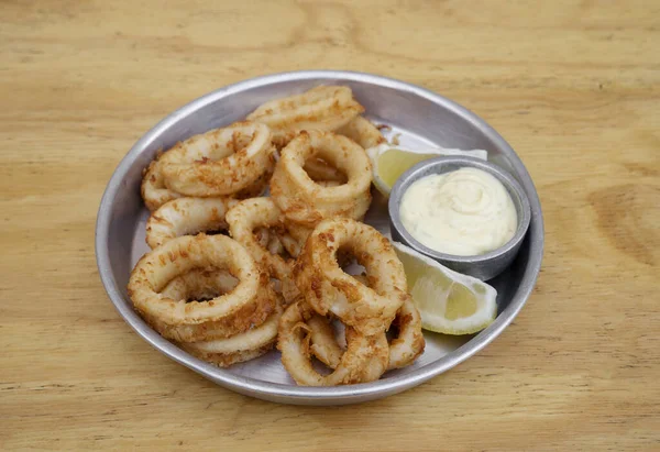 stock image Seafood. Closeup view of fried squid rings with lemon and a dipping sauce in a metal dish on the wooden table.