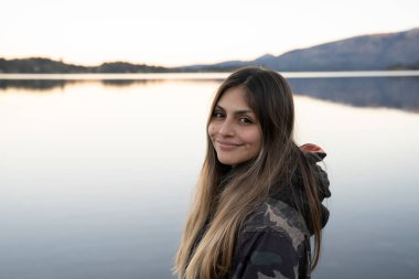 Gülümseyen genç bir kadının portresi. Arka planda durgun göl ve sıradağlar var. Çevrede güzel günbatımı renkleri.