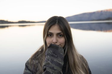 Gülümseyen genç bir kadının portresi. Arka planda durgun göl ve sıradağlar var. Güzel günbatımı renkleri. 